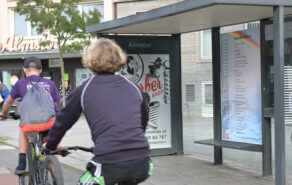 Ein LeseZeichen an der Bushaltestelle Almstor. Zwei Fahrradfahrer fahren im Vordergrund vorbei.