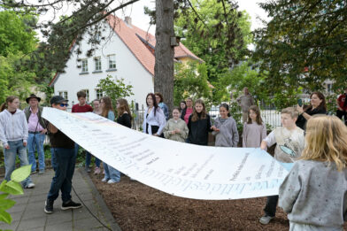 Die Schüler:innen stehen mit Initiator Jo Köhler und Lehrerin Marianne Aselmeyer zusammen und bestaunen das Lesezeichen.