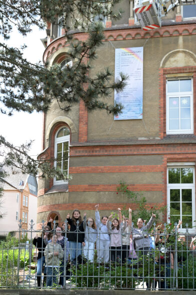 Das Lesezeichen hängt an der Schule. Die Schüler:innen stehen vor der Schule und zeigen den Daumen nach oben.
