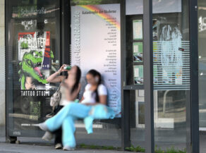 Lesezeichen an der Bushaltestelle Schuhstraße. Zwei junge Frauen sitzen vor dem Lesezeichen und trinken ein Getränk aus einer Dose.
