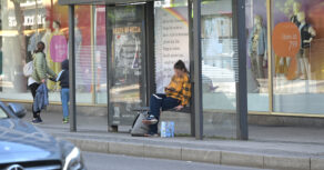 Lesezeichen an der Bushaltestelle Schuhstraße. Eine Frau sitzt vor dem Text und schaut auf ihr Handy.